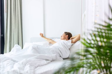 Kadın uyanıyor ve yatak odasındaki yatakta geriniyor. Beyaz iç mekan