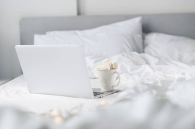 Laptopta sıcak çikolatanın üstüne tamamen marşmelov konmuş ve beyaz yatağa yerleştirilmiş beyaz bir Noel kupası. Uzayı kopyala Bayram kutlaması konsepti. Günaydın sıcak kahvaltı..