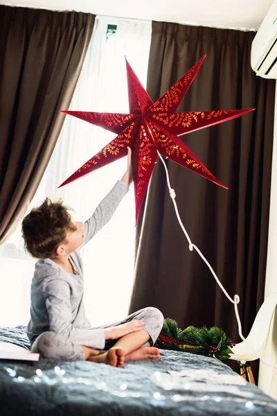男の子は赤い星でクリスマスのために家と窓を飾る 冬休みの魔法 — ストック写真