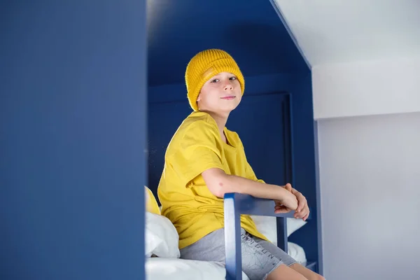 黄色のTシャツと帽子の男の子は家で青の二段ベッドの上にガウンを座って時間を費やしています — ストック写真