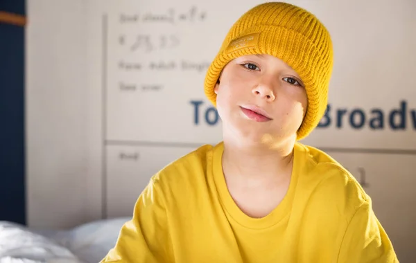 Boy in yellow t-shirt and hat is spending time sitting gown on a blue two-tier bunk bed at home.