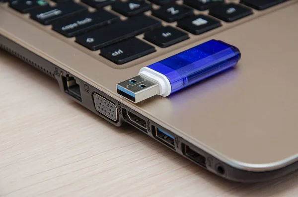 Blue USB flash drive connected to laptop computer\'s USB port. Notebook is on a light gray desk background.