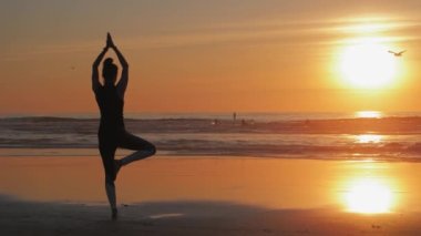 Kolları havada ince bir kadın silueti, sahilde yoga pratiği.