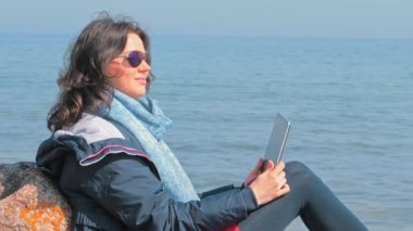 Kaya sahilinde dizüstü bilgisayarıyla oturan ve bahar güneşinin tadını çıkaran bir kadın.