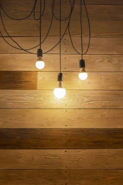 Uzun siyah kablolar üzerinde üç modern LED lambadan yapılmış bir lamba ahşap bir duvarın karşısında. Ahşap arka planda retro lambaları kullanarak modern stil aydınlatma