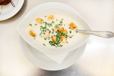 Kremalı çorba, kızarmış ekmek ve bir restorandaki masada beyaz bir tabakta otlar. Karantina sonrası gastro turizmi. Masada beyaz masa örtüsü olan soğan kremalı çorba. Evde yemek pişirmek karantina sırasında lezzetli ve sağlıklıdır.