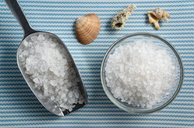 Metalik kepçe ve deniz tuzu dolu cam kase beyaz ve mavi deniz kabuklarıyla kaplı.