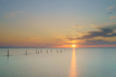 Güneş ufuktan aşağı IJsselmeer 'in sakin sularına batar ve altın bir yol açar. Ahşap direkler, bazıları ağlı, sakin denizi noktalar, sakin ve atmosferik bir deniz manzarası yaratır.