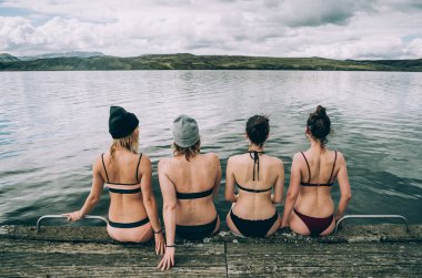 Bikini giyen bir grup kız, soğuk gölün kıyısında, İzlanda 'nın bulutlu gökyüzünün altında oturuyorlar ve uzaklara bakıyorlar.