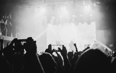 Bir konser izleyicisinin siyah beyaz fotoğrafı, kaldırılmış eller, akıllı telefonlar kayıt, sahne ışıkları, ve enerjik bir enerji ve özgürlük atmosferi..