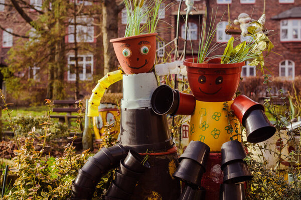 Creative garden decoration in Nikiszowiec, Katowice, Poland, featuring two human-like figures made from colorful plastic flower pots, with green plants as hair and cheerful painted faces.