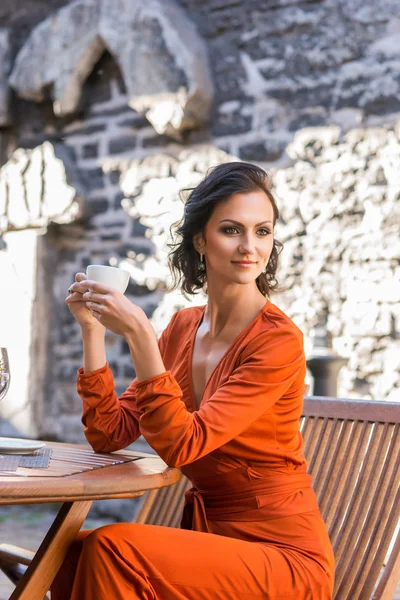 Vacker elegant kvinna i orange mantel dricka kaffe på terrassen på restaurang — Stockfoto