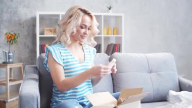 Genç ve mutlu bir kadın evdeki kanepede oturan paket kutusunu açıyor. Teslimat konsepti.