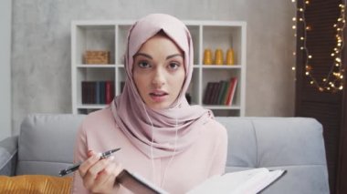 Genç ve çekici tesettürlü Müslüman kadın evde kulaklıkla çalışıyor. Orta Doğulu serbest çalışan ya da öğrenci çevrimiçi konuşma yapıyor. Kilitleme kavramı.