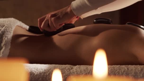 Mujer joven y hermosa recibe terapia de masaje de piedra caliente en el salón de spa. Estilo de vida saludable y concepto de cuidado corporal. — Vídeos de Stock