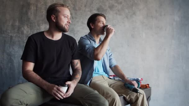 Twee vrienden rusten en praten tijdens een pauze van de interieurrenovatie. Mannelijke werknemers zitten op steigers en drinken koffie. — Stockvideo