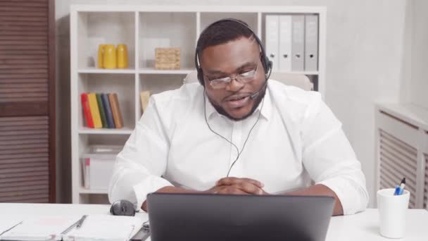 フリーランスの職場。アフリカ系アメリカ人の男性は、コンピュータとヘッドセットを使って自宅のオフィスで働いています。従業員が電話会議を開いている。リモートジョブコンセプト. — ストック動画