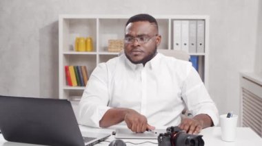 Ev ofisinde serbest fotoğrafçı işyeri. Genç Afro-Amerikalı adam bilgisayar, grafik tablet ve kamera kullanıyor. Uzak görev konsepti.