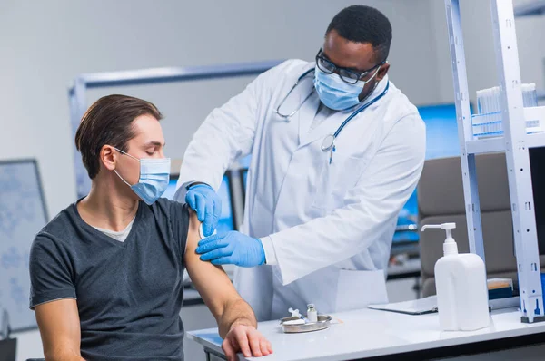 O médico afro-americano faz uma vacina contra o coronavírus usando uma seringa e uma agulha hipodérmica. Médico profissional e paciente no consultório do hospital. Vacinação e segurança. — Fotografia de Stock