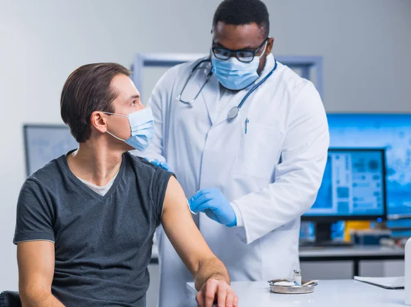 O médico afro-americano faz uma vacina contra o coronavírus usando uma seringa e uma agulha hipodérmica. Médico profissional e paciente no consultório do hospital. Vacinação e segurança. — Fotografia de Stock