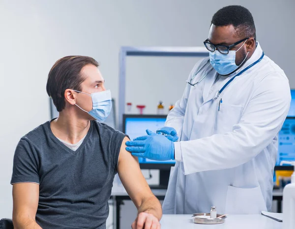 O médico afro-americano faz uma vacina contra o coronavírus usando uma seringa e uma agulha hipodérmica. Médico profissional e paciente no consultório do hospital. Vacinação e segurança. — Fotografia de Stock