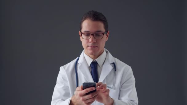 Retrato de estúdio de jovem médico profissional usando dispositivo de smartphone sobre fundo cinza — Vídeo de Stock