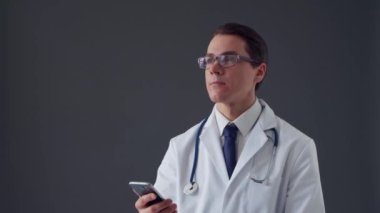 Gri arkaplan üzerinde akıllı telefon cihazı kullanan genç profesyonel tıp doktorunun stüdyo portresi