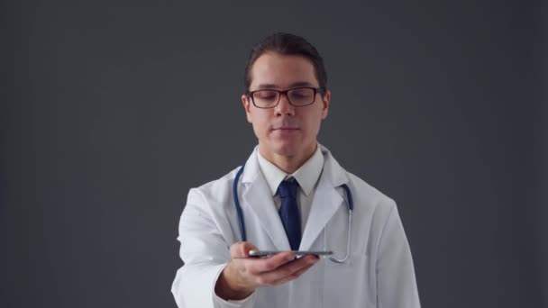 灰色背景下使用智能手机装置的年轻专业医生的工作室肖像 — 图库视频影像