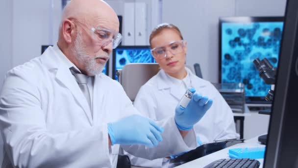 Profesor de edad avanzada y joven asistente de laboratorio están haciendo experimentos científicos en un laboratorio moderno. Laboratorio de ingenieros genéticos. El concepto de ciencia, medicina y desarrollo de vacunas. — Vídeos de Stock