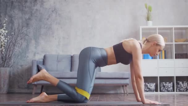 Menina jovem e desportiva em sportswear está fazendo exercícios em casa interior usando banda de resistência. Fit e mulher loira esbelta entra para esportes e fitness. Saúde, queima de gordura e bem-estar. — Vídeo de Stock