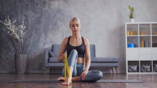 Menina jovem e desportiva em sportswear está fazendo exercícios em casa interior usando banda de resistência. Fit e mulher loira esbelta entra para esportes e fitness. Saúde, queima de gordura e bem-estar. — Vídeo de Stock