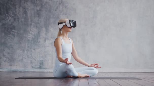 Chica joven y deportiva en ropa deportiva está haciendo ejercicios de yoga utilizando casco de realidad virtual. Ajuste y delgada mujer rubia entra en los deportes y la aptitud. Salud, quemaduras de grasa y. — Vídeo de stock