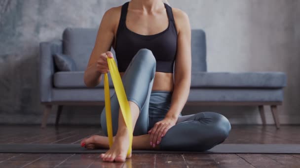 Giovane e sportiva ragazza in abbigliamento sportivo sta facendo esercizi in casa interna utilizzando banda di resistenza. In forma e snella donna bionda entra per lo sport e il fitness. Assistenza sanitaria, bruciare i grassi e benessere. — Video Stock