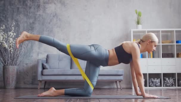 Menina jovem e desportiva em sportswear está fazendo exercícios em casa interior usando banda de resistência. Fit e mulher loira esbelta entra para esportes e fitness. Saúde, queima de gordura e bem-estar. — Vídeo de Stock