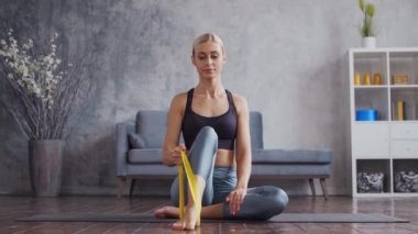 Spor giyimli genç ve sportif kız direniş bandını kullanarak iç mekanda egzersiz yapıyor. Formda ve narin sarışın kadın spor ve spor için geliyor. Sağlık sigortası, yağ yakma ve sağlık..