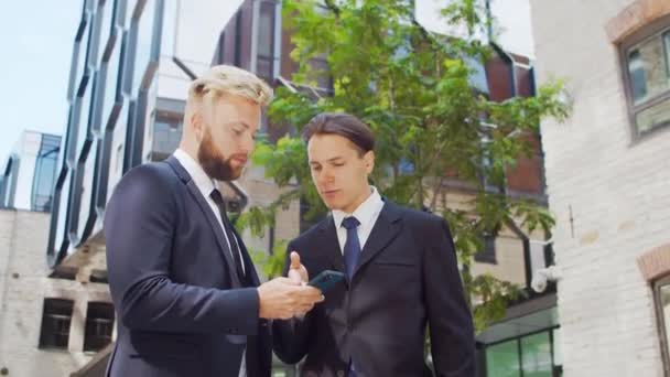 Självsäker affärsman och hans kollega framför en modern kontorsbyggnad. Finansiella investerare använder smartphone-enhet och pratar. Banker och företag. — Stockvideo