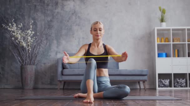 Das junge, sportliche Mädchen in Sportbekleidung macht im heimischen Innenraum Übungen mit einem Widerstandsband. Die durchtrainierte und schlanke Blondine treibt Sport und Fitness. Gesundheit, Fettverbrennung und Wellness. — Stockvideo