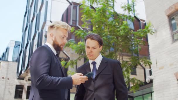 Zelfverzekerde zakenman en zijn collega tegenover modern kantoorgebouw. Financiële beleggers gebruiken smartphone en praten. Bankieren en bedrijfsleven. — Stockvideo