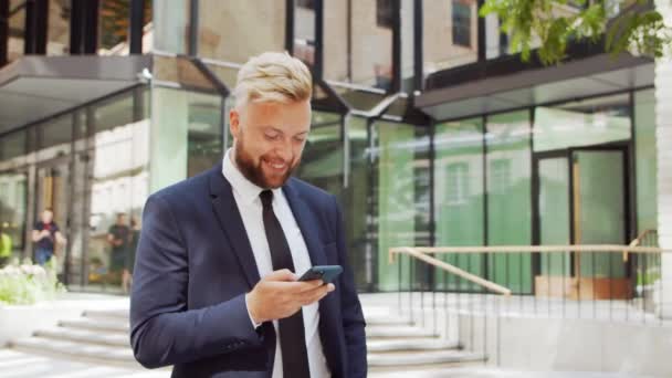 Självsäker affärsman framför en modern kontorsbyggnad. Finansiell investerare använder smartphone utomhus. Banker och företag. — Stockvideo