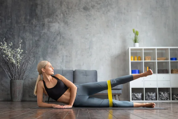 Menina jovem e desportiva em sportswear está fazendo exercícios em casa interior usando banda de resistência. Fit e mulher loira esbelta entra para esportes e fitness. Saúde, queima de gordura e bem-estar. — Fotografia de Stock