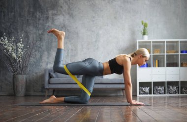 Spor giyimli genç ve sportif kız direniş bandını kullanarak iç mekanda egzersiz yapıyor. Formda ve narin sarışın kadın spor ve spor için geliyor. Sağlık sigortası, yağ yakma ve sağlık..