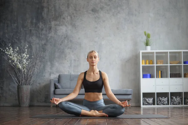 Chica joven y deportiva en ropa deportiva está haciendo ejercicios de yoga en el interior del hogar. Ajuste y delgada mujer rubia entra en los deportes y la aptitud. Salud, quemaduras de grasa y bienestar. — Foto de Stock