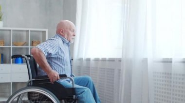 Engelli yaşlı adam evde tek başına tekerlekli sandalyede oturuyor. Tekerlekli sandalyedeki engelli pencereden dışarı bakıyor. Felç ve sakatlık.