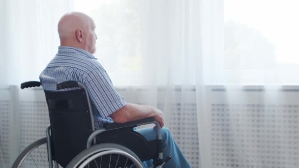 El viejo discapacitado está sentado en una silla de ruedas en casa solo. Una persona discapacitada en silla de ruedas está mirando por la ventana. Parálisis y discapacidad. — Vídeos de Stock