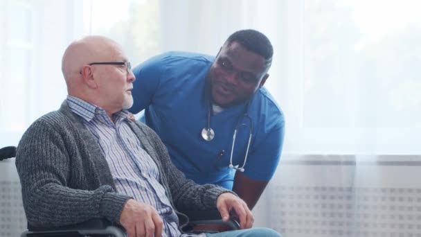 African-American caregiver and old disabled man in a wheelchair. Professional nurse and handicapped patient in a nursing home. Assistance, rehabilitation and health care. — Stock Video