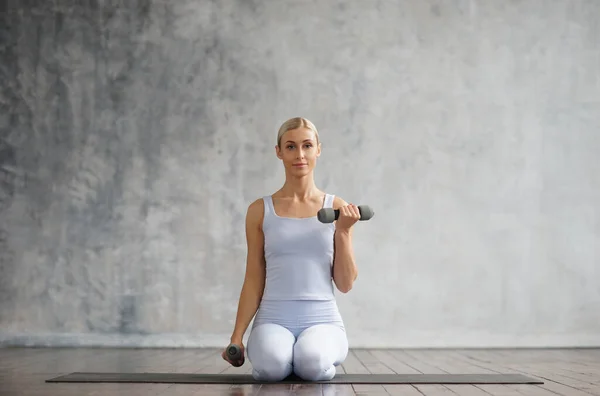 Young and sporty girl in sportswear is doing exercises in home interior. Fit and slender blond woman goes in for sports and fitness. Healthcare, fat burn and wellness. — Stock Photo, Image