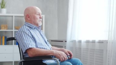 Engelli yaşlı adam evde tek başına tekerlekli sandalyede oturuyor. Tekerlekli sandalyedeki engelli pencereden dışarı bakıyor. Felç ve sakatlık.