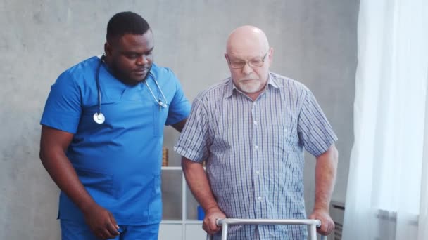 Eine afroamerikanische Pflegerin bringt einem behinderten alten Mann das Gehen mit Rollator bei. Fachkrankenschwester und behinderte Patientin in einem Pflegeheim. Hilfe, Rehabilitation und Gesundheitsfürsorge. — Stockvideo