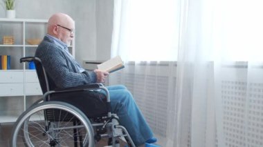 Engelli yaşlı adam evde tek başına tekerlekli sandalyede oturuyor. Tekerlekli sandalyedeki bir özürlü kitabı okuyor. Felç ve sakatlık.