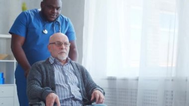 Afro-Amerikalı bakıcı ve tekerlekli sandalyedeki yaşlı engelli adam. Bakımevindeki profesyonel hemşire ve özürlü hasta. Yardım, rehabilitasyon ve sağlık hizmetleri.